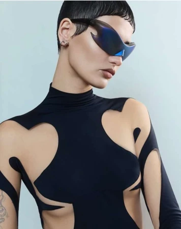 Model with short dark hair wearing futuristic blue mirrored sunglasses and a black cut out top against a light background