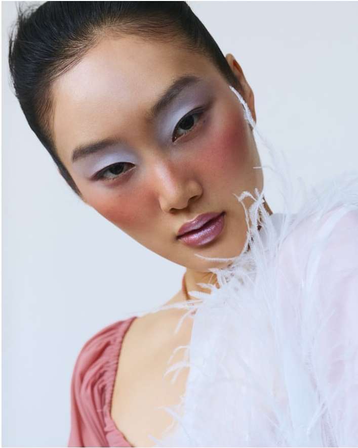 Close-up portrait of an East Asian woman with lavender eyeshadow, pink blush, and glossy lips wearing a feather-trimmed white garment