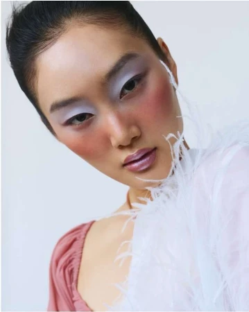 Close-up portrait of an East Asian woman with lavender eyeshadow, pink blush, and glossy lips wearing a feather-trimmed white garment