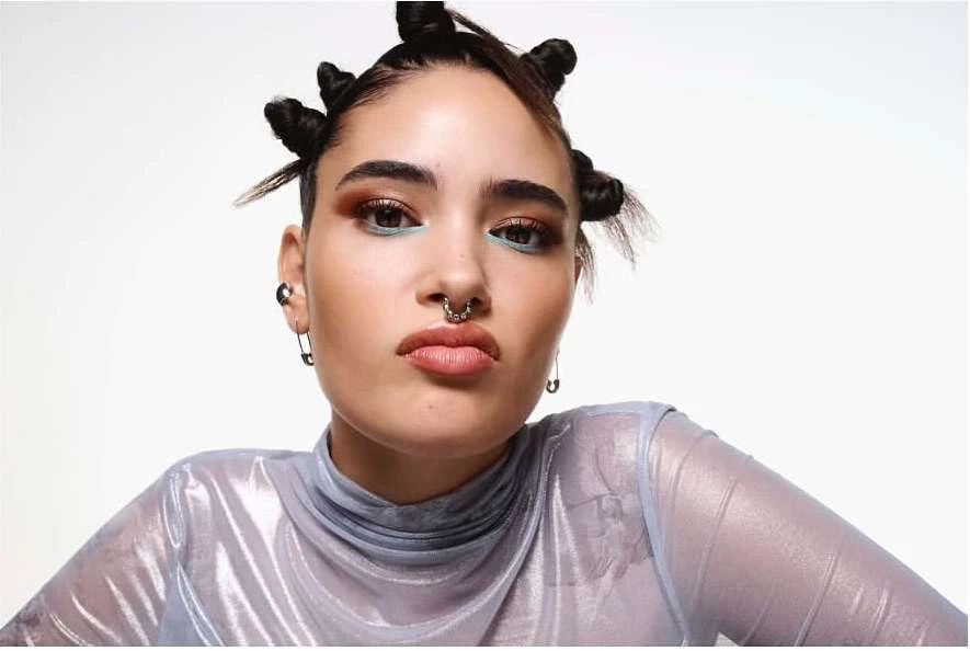 Close-up of a young woman with space buns hairstyle and a silver shimmering top against a white background in a beauty studio shot.