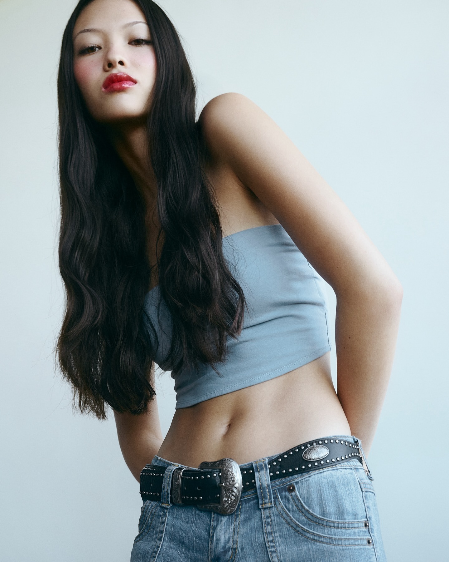 Portrait of a young adult Asian woman posing in a tube top and jeans with a western belt, set against a light neutral background indoors.