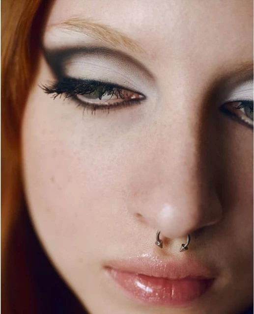 Close-up portrait of a young woman with fair skin, red hair, nose ring, and smokey eye makeup, focusing on beauty and personal expression.
