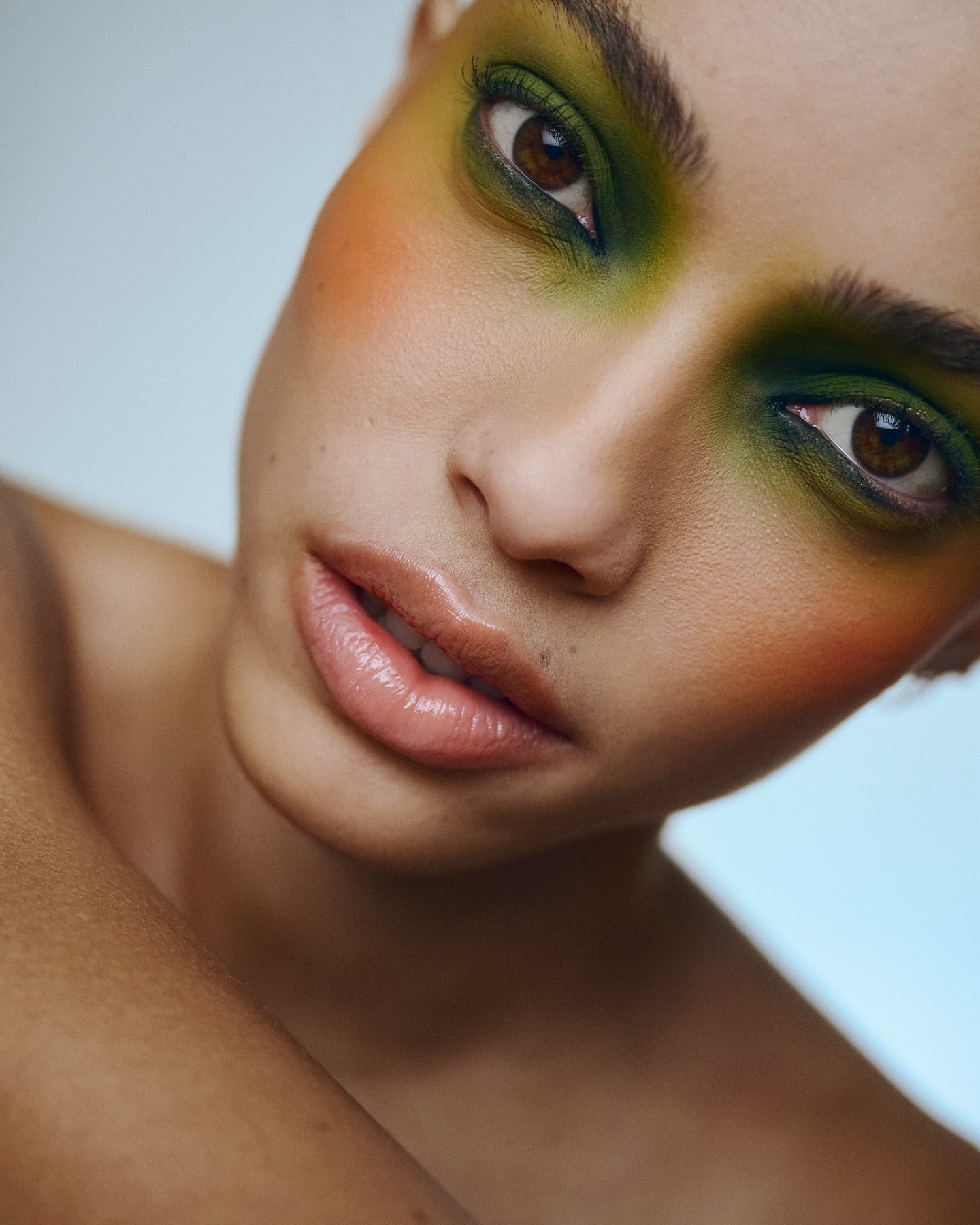 Close-up portrait of a woman with green eyeshadow, peach blush, and glossy lips, set against a light blue backdrop in a beauty editorial.