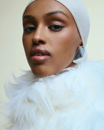 Close-up portrait of a woman wearing a white cap and fluffy collar with silver earrings against a plain background