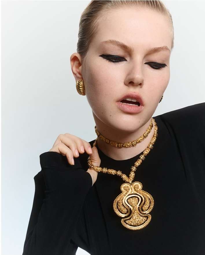 Close-up of a young woman with heavy black eyeliner wearing a black shirt and gold statement jewelry against a white backdrop.