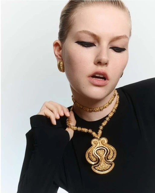Close-up of a young woman with heavy black eyeliner wearing a black shirt and gold statement jewelry against a white backdrop.
