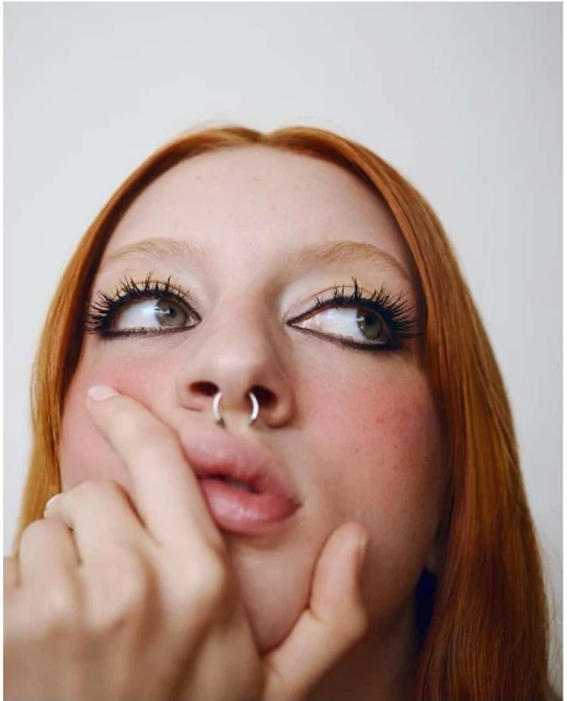 Portrait of a red-haired woman with a nose ring looking up and to the side, with heavy black eyeliner and pink blush, against white.