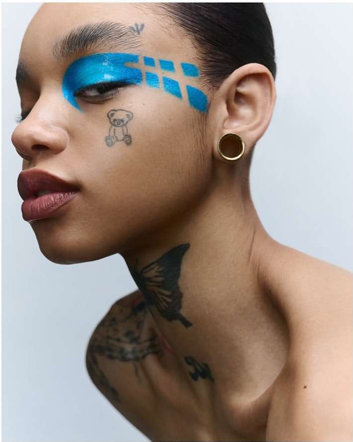 Close-up portrait of a woman with creative blue eyeshadow and face tattoos, emphasizing beauty and body art against a plain background.