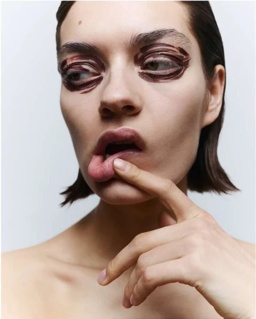 Close-up portrait of a young fair-skinned woman with dramatic brown eye makeup, finger on lip, fair skin, and short brown hair.