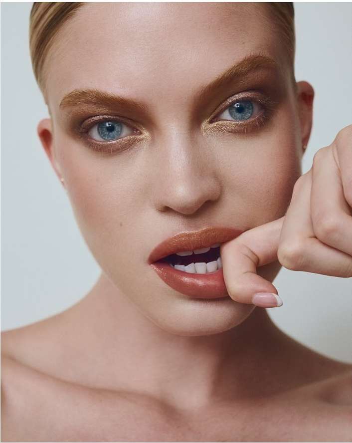 Portrait of a fair-skinned woman with striking blue eyes and warm-toned makeup, biting her finger in a close-up studio shot.