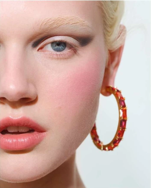 Close-up portrait of an adult female model with pale skin, wearing bright eye makeup, an earring, and looking at the camera.