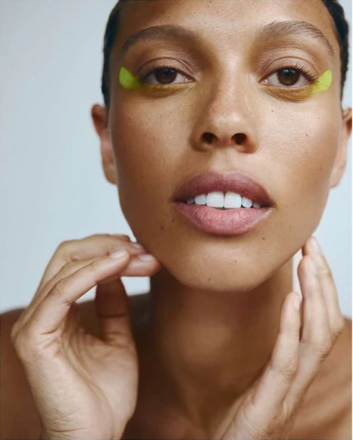 Close-up portrait of an adult female with fair skin, light pink lips, and green under-eye patches, hands gently touching her face.