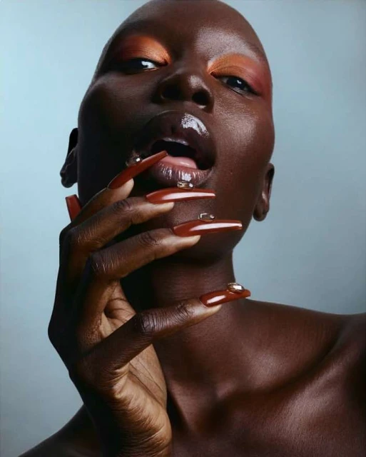 Close-up studio portrait of a Black female model with orange eye makeup, long maroon nails, and glossy lips with water droplet accents.