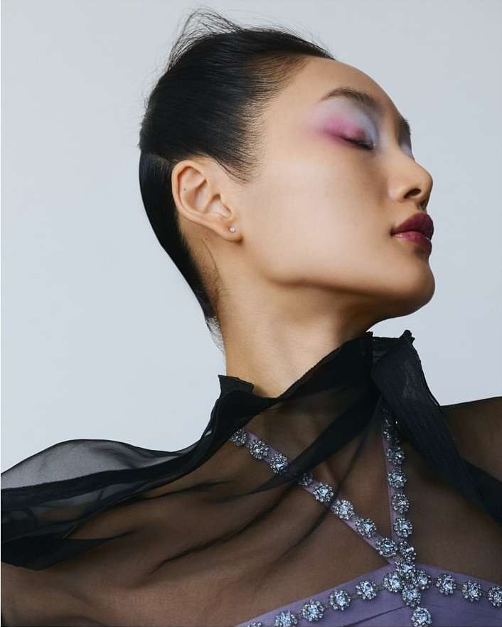 Profile of Asian woman with eyes closed wearing purple dress and sheer black cape, face highlighted with bright pink and purple eyeshadow.