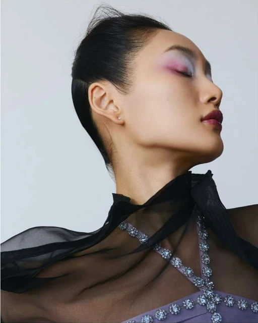 Profile of Asian woman with eyes closed wearing purple dress and sheer black cape, face highlighted with bright pink and purple eyeshadow.