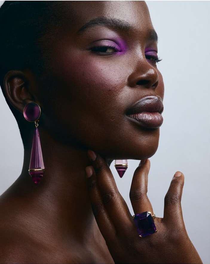 Close-up portrait of a Black woman with purple eyeshadow and jewellery, including earrings and a ring, against a neutral background.