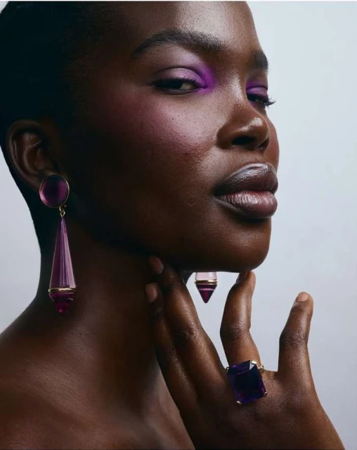 Close-up portrait of a Black woman with purple eyeshadow and jewellery, including earrings and a ring, against a neutral background.