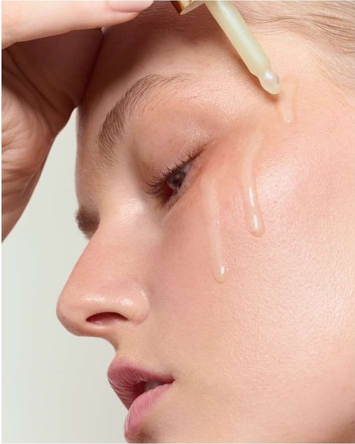 Close-up of a fair-skinned adult woman applying serum from a dropper to her face in a beauty shot, showing clear skincare and hydration.
