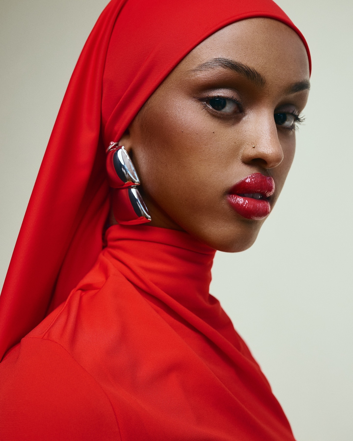 Close-up portrait of an adult female wearing a bright red headscarf, matching high-neck top, and bold silver earrings.