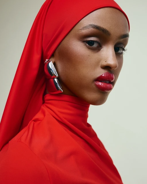 Close-up portrait of an adult female wearing a bright red headscarf, matching high-neck top, and bold silver earrings.