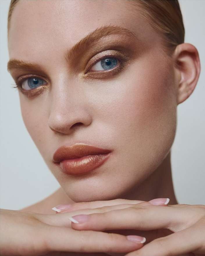 Close-up portrait of an adult female with bronze eyeshadow, glossy lips, and manicured nails, resting her chin on her interlocked hands.