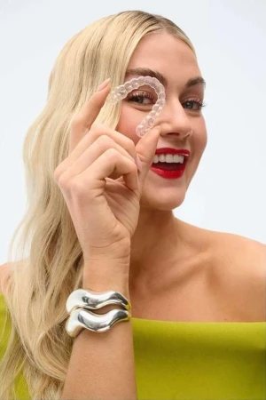 Woman smiles, holding a clear dental aligner by her eye, promoting dental care and orthodontic solutions for teeth straightening.