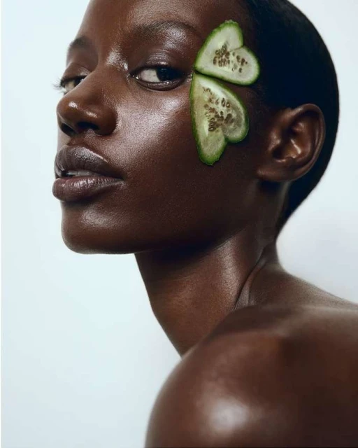 Striking close-up of a dark-skinned woman with cucumber slices near her eye against a clean white backdrop, highlighting skincare.