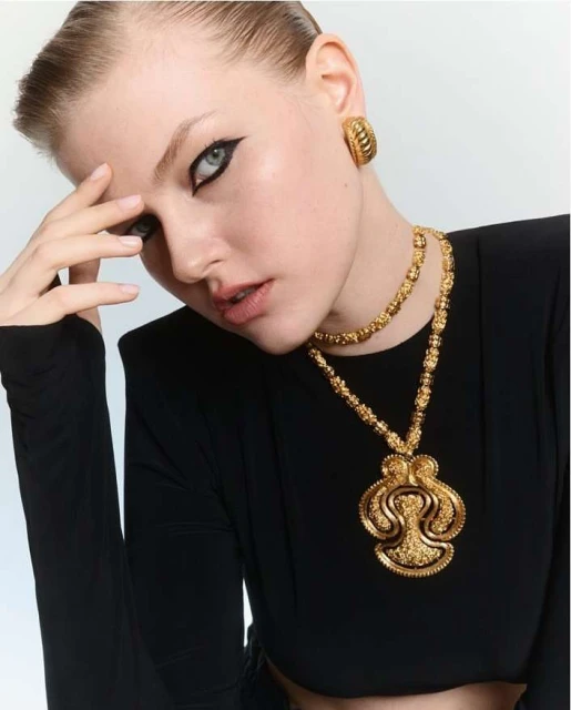 Close-up shot of a young woman with dramatic eye makeup, wearing gold statement jewelry and a black top, against a bright background.