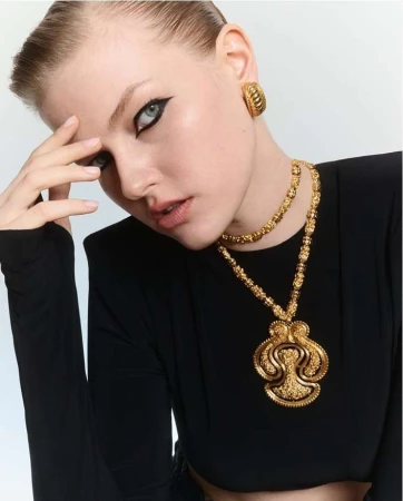 Close-up shot of a young woman with dramatic eye makeup, wearing gold statement jewelry and a black top, against a bright background.