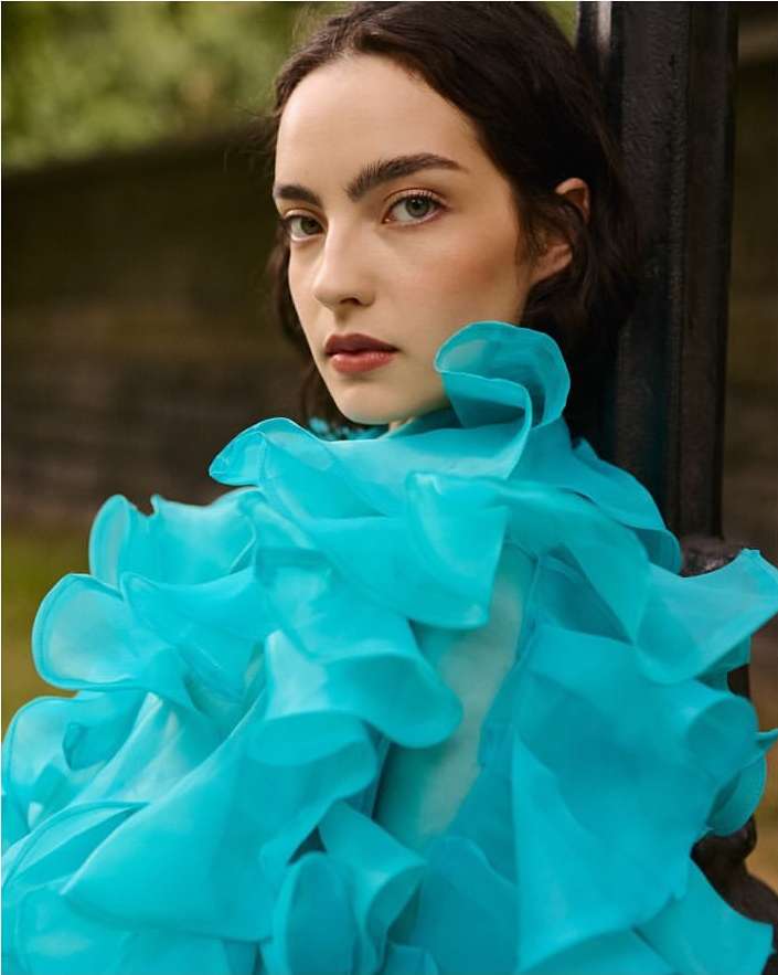 Portrait of a young woman with dark hair and a light complexion wearing a large, ruffled bright turquoise top against a dark backdrop.