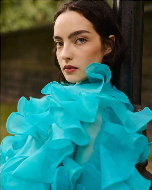Portrait of a young woman with dark hair and a light complexion wearing a large, ruffled bright turquoise top against a dark backdrop.