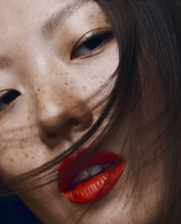 Close-up portrait of an East Asian woman with visible freckles and bright red lipstick, framed by loose dark hair strands across her face.