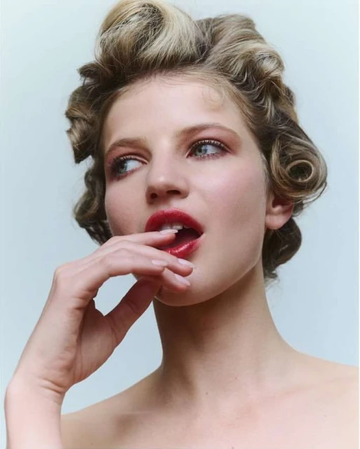 Close-up portrait of a young adult woman with blonde curly hair and red lipstick, looking to the side in a light background.