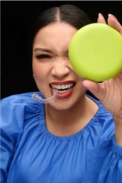 Adult female holds aligner case to her face, showing a dental aligner in her mouth, with a black background, medium shot, beauty.