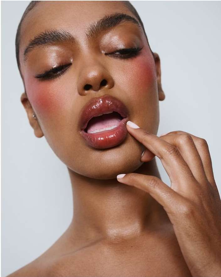 Close-up beauty shot of a dark-skinned woman with glossy lips, pink blush, and winged eyeliner against a bright white background.