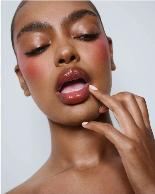 Close-up beauty shot of a dark-skinned woman with glossy lips, pink blush, and winged eyeliner against a bright white background.