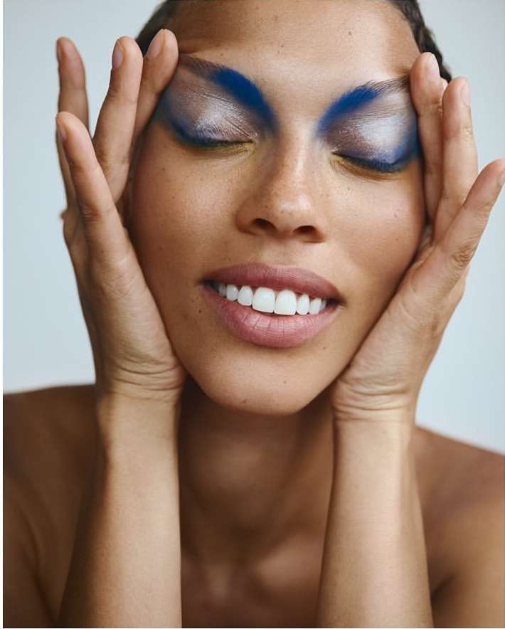 Close-up beauty shot of a smiling adult female with vibrant blue eye makeup, fair skin, and hands framing her face against a white backdrop.