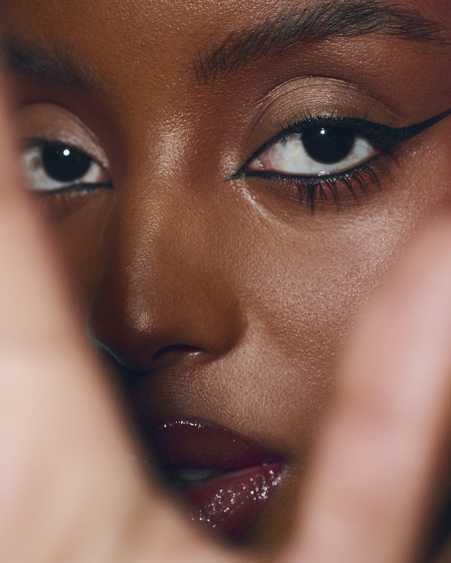 Close-up studio shot of an adult Black woman with winged eyeliner and glossy lips. The subject's fingers partially frame her face.