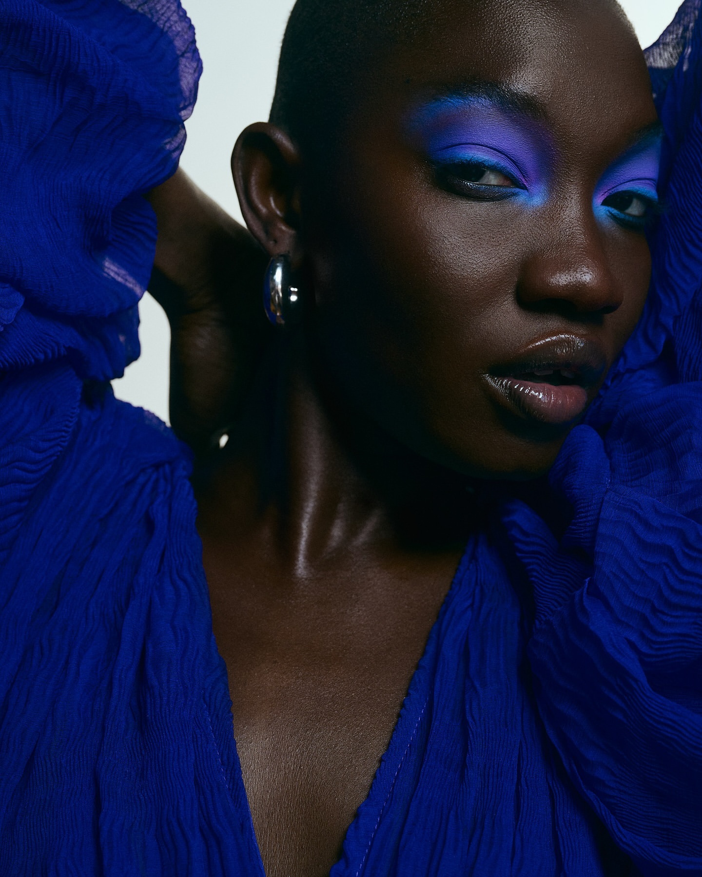 Close-up of a Black woman with a shaved head, wearing silver hoop earrings and bright blue and purple eye makeup, against a white background.