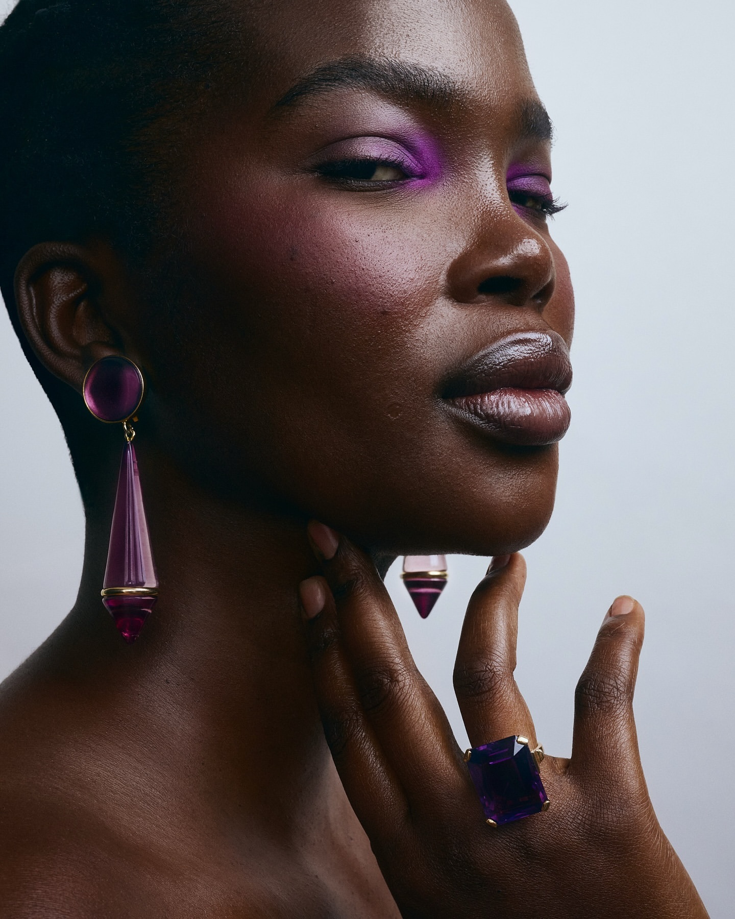 Striking close-up of a Black woman wearing purple eyeshadow and geometric amethyst jewelry, set against a soft gradient backdrop.