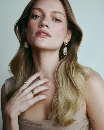 Close-up portrait of a woman with blonde hair wearing gold drop earrings and rings against a pale background