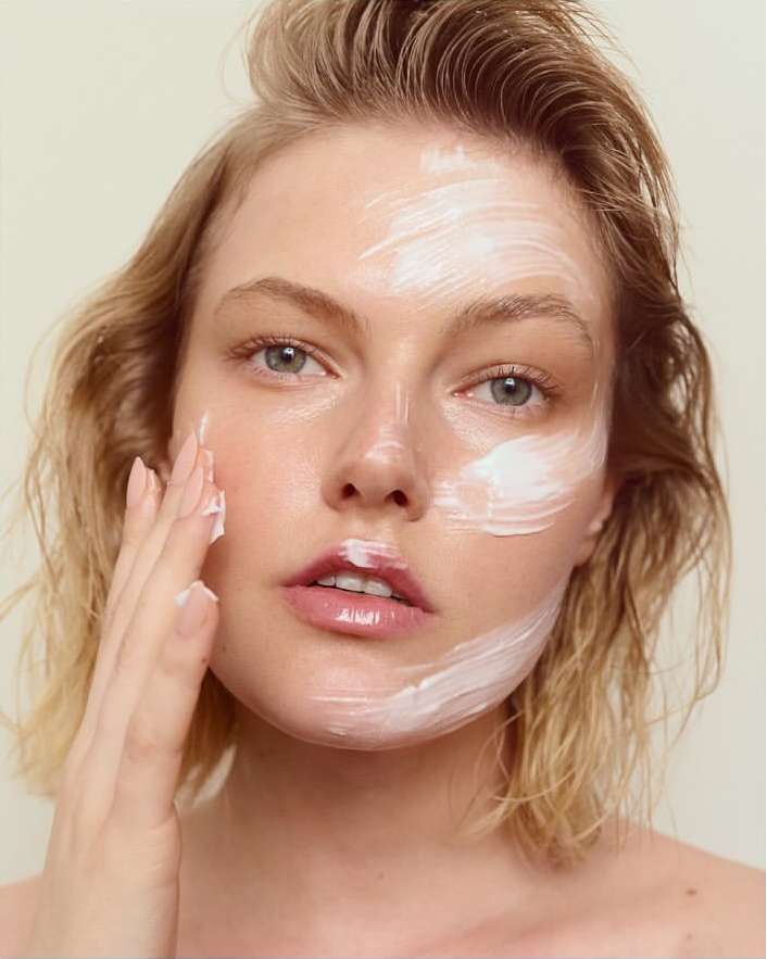 Close-up of an adult woman applying white facial cream with fingers, fair skin, blonde hair, neutral background; beauty and skincare concept.