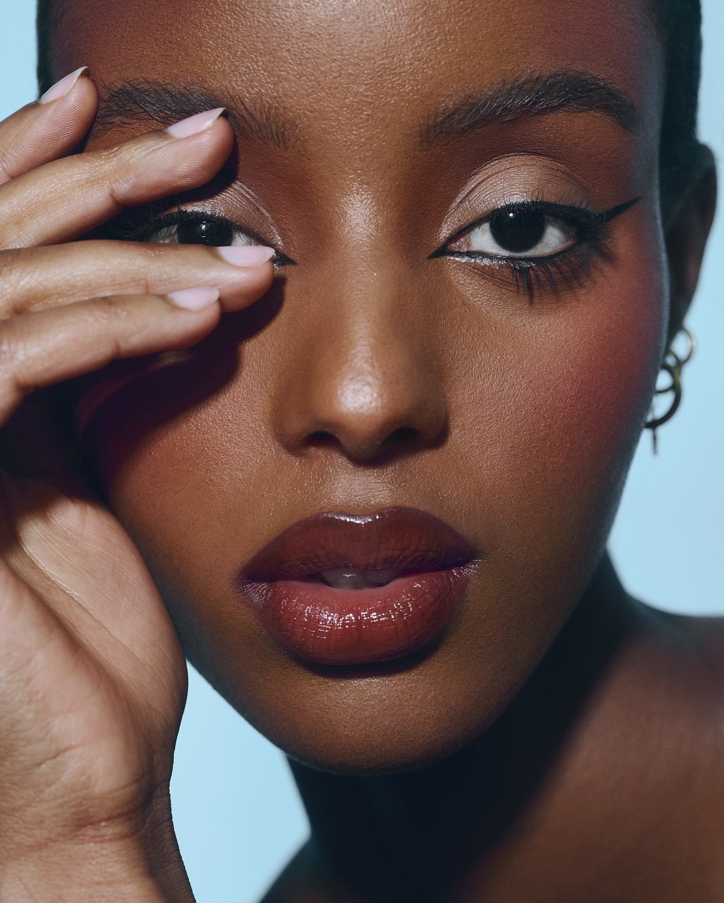 Close-up beauty shot of a dark-skinned woman with flawless makeup and glossy lips, partially obscuring her face with her hand.