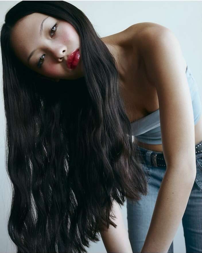 Portrait of a woman with long dark wavy hair wearing a blue crop top and denim jeans against a plain white background.