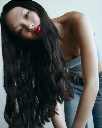 Portrait of a woman with long dark wavy hair wearing a blue crop top and denim jeans against a plain white background.