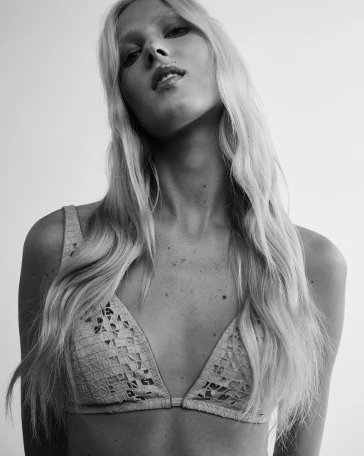 Black-and-white portrait of a fair-skinned adult woman with long blonde wavy hair, wearing a lacy bra, against a plain background.