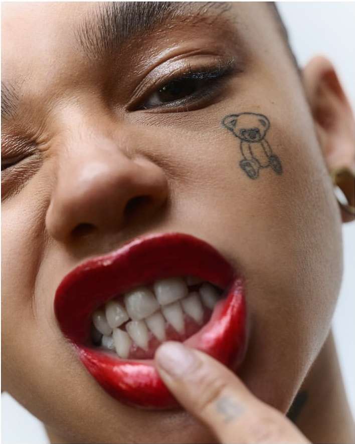Extreme close-up portrait of a person with glossy red lips, a small bear cheek tattoo, and a finger touching teeth.