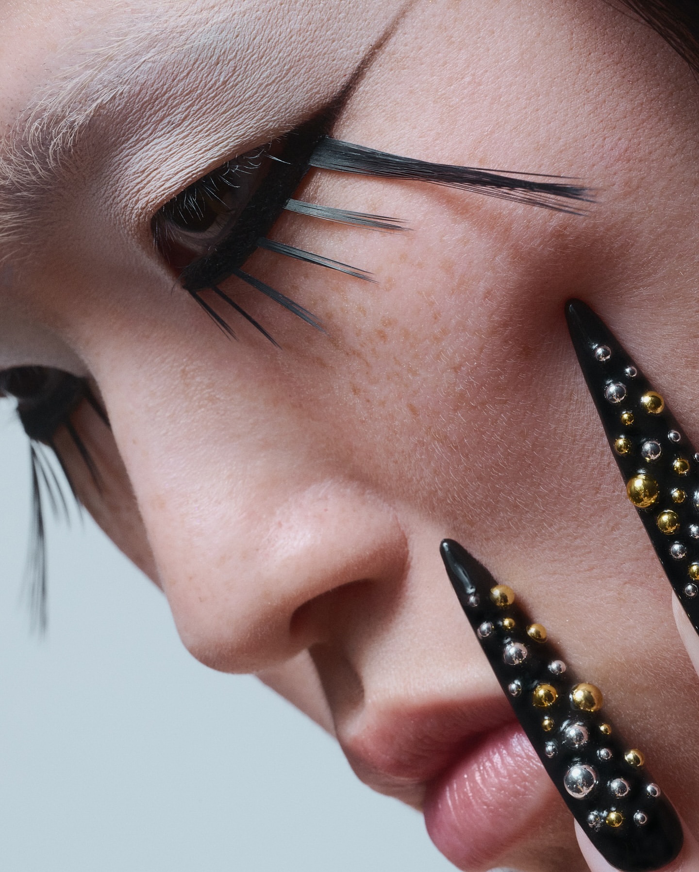 Close-up beauty shot of an adult female's face with striking feather eyelashes and bold, studded stiletto nail extensions.