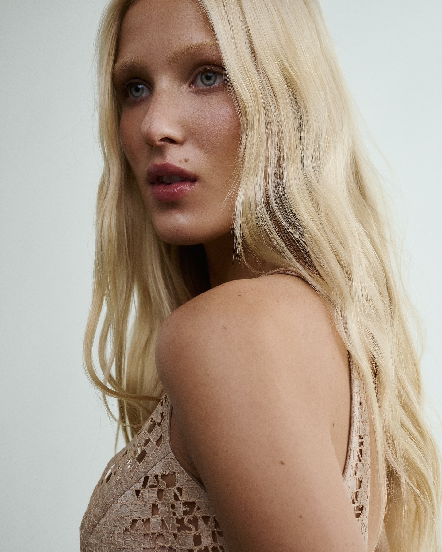 Close-up portrait of a blonde woman wearing a patterned beige textured top against a plain light wall.