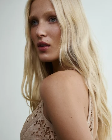 Close-up portrait of a blonde woman wearing a patterned beige textured top against a plain light wall.